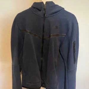 Blå zip-hoodie från Nike, storlek M - Mörkblå hoodiejacka från Nike med hel dragkedja, huva och diskret logga på bröstet. Jackan har en snygg detalj med svarta linjer över bröstet och en ficka med dragkedja på ärmen. Perfekt för chill dagar eller när du är på språng.