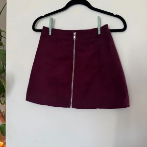 Vinröd kjol från H&M Divided - Vinröd kortkjol från H&M Divided. Kjolen har en rak passform och en snygg silverfärgad dragkedja framtill som detalj. Perfekt att styla med stickad tröja eller t-shirt för en cool look.