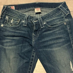 True Religion blå jeans med kontrastsömmar - Snygga blå jeans från True Religion med tydliga vita kontrastsömmar och ikoniska fickdetaljer bak. Jeansen har klassisk femficksdesign, låg midja och raka ben. Materialet är slitstarkt denim i bomull och passformen är normal. Perfekta för en avslappnad streetstil.
