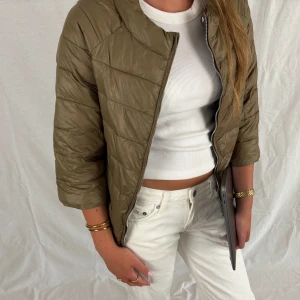 Olivgrön trekvartsärmad jacka - Snygg olivgrön pufferjacka med quiltad design och rund hals. Jackan har trekvartsärmar och stängs med dragkedja framtill. Insidan har ett prickigt foder som ger en cool kontrast. Perfekt för lager-på-lager och chill stil.
