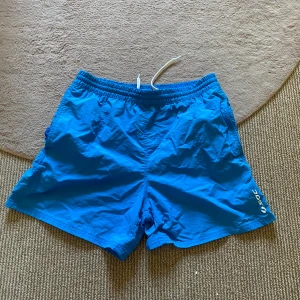 Badshorts - Snygga blå badshorts från SOC med elastisk midja och vit snörning. Shortsen har meshfoder, sidofickor och en bakficka med kardborre. Perfekta för stranden eller poolen, klassisk modell i lätt syntetmaterial🙌🏻