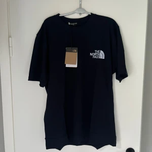 Mörkblå t-shirt från The North Face XL - Mörkblå t-shirt från The North Face i storlek XL. Klassisk modell med rund hals, korta ärmar och vit broderad logga på bröstet. Tillverkad i mjuk bomull för en skön känsla. Perfekt för en clean och stilren look.
