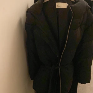 Svart pufferjacka från NLY Trend - Svart pufferjacka från NLY Trend med huva, dragkedja och bälte i midjan. Jackan har en oversized look och är perfekt för kalla dagar. Fylld med syntetmaterial för extra värme och har långa ärmar. Enkel och stilren design som funkar till allt. Trendig. Använt den få gånger, storlek 38, M. Perfekt för vintern. Den värmde verkligen gångerna jag hade på mig. Rekommenderar . 