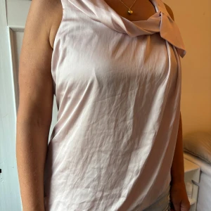 Ljusrosa ärmlös linnetopp med rosett - Supersöt ljusrosa linnetopp i slätt, glansigt material. Toppen är ärmlös och har en snygg drapering framtill med en stor rosett på ena axeln som ger en extra touch. Perfekt att styla med jeans eller kjol för en trendig look.