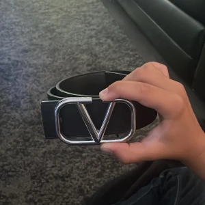 Svart Valentino bälte med silverspänne - Snyggt svart bälte från Valentino med ett stort, stilrent silverspänne i form av ett V. Bältet är gjort i äkta skinn och har en klassisk, rak form som passar till jeans eller kostymbyxor. Perfekt accessoar för att lyfta din outfit. Nästan som ny.