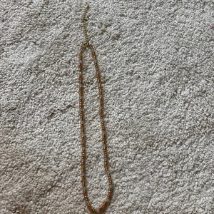 Guldigt halsband med pärldetaljer - Säljer ett tunt halsband med små guldiga pärlor och inslag av orange. Kedjan är justerbar och ger en snygg och enkel look som passar till många outfits. Perfekt accessoar för att lyfta din stil.