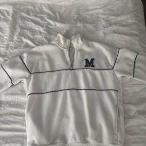 Vit half zip sweatshirt med blå detaljer - Vit sweatshirt med blå ränder och broderad M på bröstet. Tröjan har half zip-dragkedja och hög krage, samt blå kontrastsömmar längs ärmar och bröst. Perfekt för en sportig och avslappnad look.
