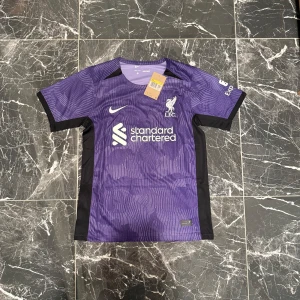 Lila Liverpool Diogo J. Nike tröja S - Liverpool FC fotbollströja i lila med svarta detaljer, officiell Nike-design. Tryck med Diogo J. och nummer 20 på ryggen. Standard Chartered-logga på bröstet. Kortärmad och tillverkad i lätt, ventilerande material.
