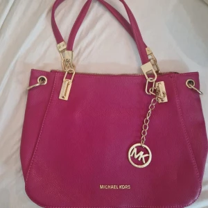 Rosa axelväska från Michael Kors - Säljer en snygg rosa axelväska från Michael Kors med guldfärgade metalldetaljer och MK-hänge. Väskan är i skinn och har dubbla handtag samt dragkedja upptill. Perfekt för dig som vill ha en färgklick till outfiten!
