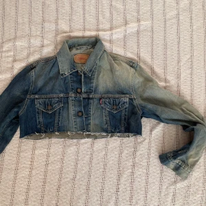 Croppad jeansjacka från Levi's  - Säljer en croppad jeansjacka från Levi's i blå denim med coolt tryck på ryggen. Jackan har knappar framtill, två bröstfickor och rå, fransig nederkant. Perfekt för dig som vill sticka ut med en unik och edgy stil.