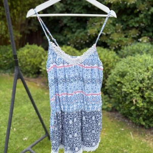 Blå mönstrad byxdress Stories - Supersöt byxdress från Stories med tunna axelband och v-ringning. Den har ett blått och vitt mönster med inslag av rött, spetsdetaljer vid hals och nederkant samt flowig passform. Perfekt för varma dagar och ger en chill boho-vibe.