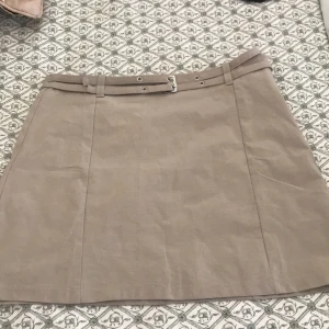 Beige kortkjol med bälte från MNG - Supersnygg beige kortkjol från MNG med dubbla bälten i midjan och silvriga detaljer. Kjolen har en stilren och enkel design som är lätt att matcha. Perfekt för dig som gillar minimalistisk stil och vill ha något som sticker ut lite extra.