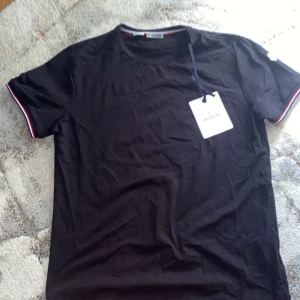 Svart Moncler t-shirt med rödvit kant - Svart moncler t-shirt Ärmarna är korta och har snygga rödvita detaljer vid mudden. Sedan Moncler-logga på ärmen och rund halsringning. Perfekt för dig som gillar stilrena och exklusiva plagg. Vid frågor skriv till mig kan mötas eller frakta priset är ej hugget i sten storlek L  plaget är helt ny aldrig använd.