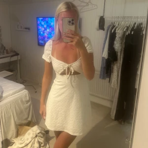 Vit kort klänning med knyt och broderi - Superfin vit kort klänning med broderade detaljer och knyt framtill. Klänningen har korta puffärmar, v-ringning och en snygg cut-out under bysten🥰