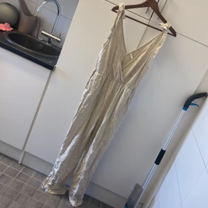 Beige byxdress från  NLY Trend - Lång, beige byxdress från NLY Trend med knytbara axelband och djup v-ringning. Tillverkad i ett lätt linneliknande material som ger en avslappnad vibe. Perfekt för varma dagar när du vill vara både bekväm och snygg. Praktisk med två fickor. 