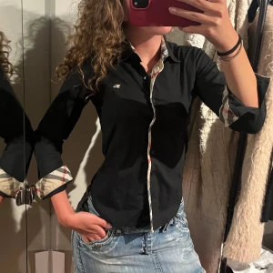 Svart Burberry skjorta  - Snygg svart skjorta från Burberry London med klassiska rutiga detaljer på insidan av kragen och ärmsluten. Skjortan har figurnära passform. Liten fläck längst ner men går bort i tvätten💞