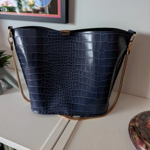 Mörkblå axelväska med kedja - Snygg mörkblå axelväska i croco-mönstrat skinnimitation med guldfärgad kedjerem och detaljer. Väskan har en stilren, något böjd form och stängs med ett litet guldfärgat spänne upptill. Perfekt för dig som gillar trendiga accessoarer med lyxig känsla. Handsydd av mig