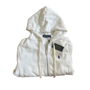 Ralph Lauren Hoodie  - Helt ny Vit hoodie från Polo Ralph Lauren med dragkedja, huva och snörning. Klassisk broderad logga på bröstet och ribbade muddar. Tillverkad i mjuk bomull, perfekt för en clean och stilren look. Längd 64cm. Jag anser att den är ganska liten så kolla måtten !