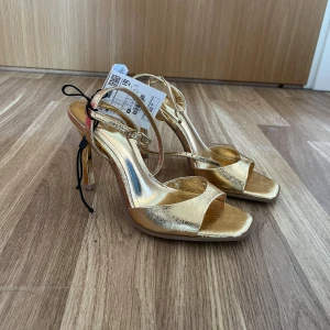 Guldiga sandaler med klack från Zara - Strappy sandaler i metallic guld från Zara med fyrkantig tå och hög klack. Skorna har smala remmar och spänne runt vristen för extra support. Perfekta för dig som vill sticka ut med en glansig och trendig look.