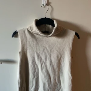 Beige stickad topp från Zara - Säljer en beige stickad väst från Zara med hög, ribbad polokrage. Västen är ärmlös och har en clean, minimalistisk look som är lätt att styla. Perfekt för lager-på-lager och passar till både jeans och kjol.