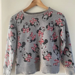 Grå Minnie Mouse tröja med prickigt mönster - Superfin grå sweatshirt med all-over print av Minnie Mouse och röda prickiga rosetter. Tröjan har rund hals, långa ärmar och ribbade muddar. Perfekt för dig som älskar Disney och vill ha en bekväm och snygg vardagströja.