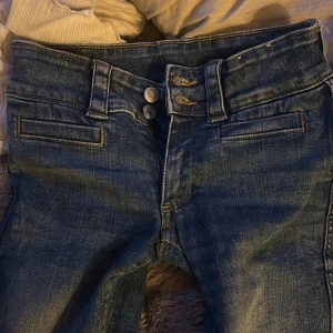 Blå bootcut jeans med dubbla knappar - Snygga blå jeans med bootcut passform och dubbla knappar i midjan. Jeansen har slitna detaljer, fickor med lock bak och dekorativa fickor fram. Perfekta för dig som gillar en klassisk men ändå trendig look.