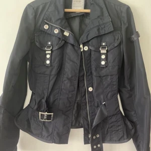 Svart Peuterey jacka med bälte och detaljer - Svart Peuterey jacka med coola spännen och fickor framtill, dragkedja och bälte i midjan. Jackan har långa ärmar och silvriga knappar som ger en edgy look. Perfekt för dig som gillar streetstyle och vill sticka ut.