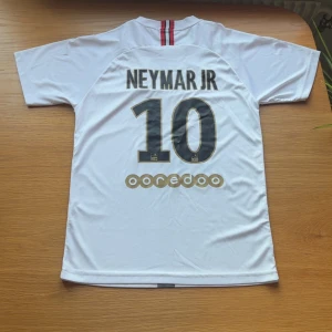 PSG Neymar Jr 10 vit matchtröja - Snygg vit PSG fotbollströja med Neymar Jr och nummer 10 på ryggen. Framsidan har Fly Emirates-tryck, PSG-märke och Jordan-logga. Tunn och lätt polyester, kortärmad med svart/grå vertikal rand. Perfekt för dig som älskar fotboll och stilren design.