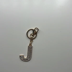 Nyckelring med glittrig bokstav J - Snygg nyckelring i guld med bokstaven J täckt av glittrande stenar på ena sidan och blank guldfinish på andra. Perfekt accessoar till väskan eller nycklarna för dig som vill sticka ut med lite extra bling.