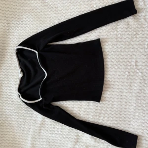 Svart långärmad offshoulder topp - Snygg svart långärmad offshoulder topp med vit kant längs halsringningen. Tröjan har en figurnära passform och är tillverkad i ett ribbat material som ger en stilren och modern look. Perfekt att styla med jeans eller kjol för en trendig outfit.