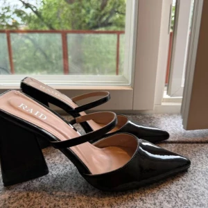 Svarta klackskor från RAID - Snygga svarta klackskor från RAID med spetsig tå och två tunna remmar över foten. Skorna har hög blockklack och en glansig finish som ger en trendig look. Perfekta för dig som vill sticka ut med stilrena detaljer.