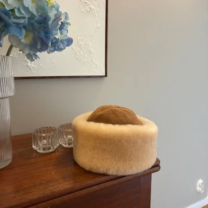 Beige pälsmössa med mockadetaljer - Snygg beige pälsmössa med bred, fluffig kant och ovandel i mjuk mocka. Klassisk och varm modell som sticker ut med sin runda form och lyxiga känsla. Perfekt för kalla dagar när du vill ha både stil och värme. Varan är helt ny och aldrig använd. Nypris 2400 kr.