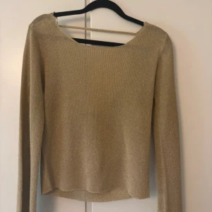 Glittrig guldig topp från Gina Tricot - Snygg glittrig guldig topp från Gina Tricot i ribbat material. Långärmad med öppen rygg och en smal rem över axlarna. Perfekt för dig som vill sticka ut med en trendig och elegant look. Modellen är figurnära och passar till många olika stilar.