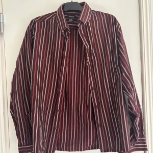 Randig skjorta från Madison - Snygg skjorta från Madison med vertikala ränder i vinrött, vitt och svart. Klassisk krage, knäppning framtill och långa ärmar. Perfekt för dig som gillar en stilren look med lite edge.