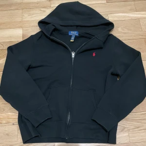 Svart hoodie från Polo Ralph Lauren - Svart hoodie från Polo Ralph Lauren med dragkedja och huva. Klassisk design med röd broderad logga på bröstet och två fickor framtill. Perfekt för en chill och stilren look. Tillverkad i mjukt bomullsmaterial för extra komfort. Använd 5 gånger bara ny skick köpt för 1200 i storlek 14-16