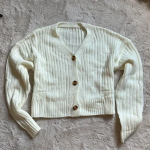 Vit ribbad kofta med knappar - Superfin vit kofta med ribbat mönster och tre stora träknappar framtill. Koftan har v-ringning och långa ärmar, perfekt att slänga på sig över en t-shirt eller linne. Croppad modell som ger en trendig look.