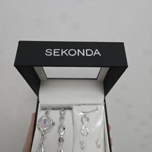 Sekonda smyckeset i silver med stenar - Snyggt smyckeset från Sekonda med armband, halsband och örhängen i silverfärg. Alla delar har en elegant åttaformad design med gnistrande stenar. Klockan har pärlemor-urtavla och matchande armband. Perfekt för dig som gillar stilrena detaljer.