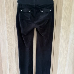 Svarta mjukisbyxor Juicy Couture XS - Svarta mjukisbyxor från Juicy Couture med bred resår och snörning i midjan. Byxorna har raka ben, stora fickor fram och bak med tryckknappar. Mjukt och glansigt material som känns lyxigt och skönt.