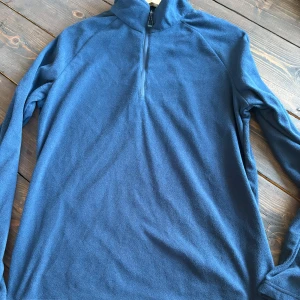 Blå fleece half zip från Mondo XL - Mysig blå fleece-tröja från Mondo i storlek XL. Tröjan har en hög krage och half zip-dragkedja framtill. Perfekt för kyliga dagar och enkel att slänga på sig över en t-shirt. Långärmad och i mjukt fleecematerial. Passar M
