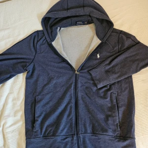 Marinblå hoodie från Polo Ralph Lauren - Snygg marinblå hoodie från Polo Ralph Lauren med dragkedja och klassisk broderad logga på bröstet. Tröjan har huva, långa ärmar och två fickor framtill. Tillverkad i mjuk bomullsmix som är skön att ha på sig hela dagen.