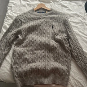 Grå kabelstickad tröja Polo Ralph Lauren - Kabelstickad grå tröja från Polo Ralph Lauren i mjuk ull. Klassisk rund halsringning och diskret broderad logga på bröstet. Perfekt för dig som gillar stilren och tidlös design med en lyxig känsla.
