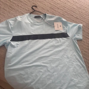 Ljusblå Emporio Armani t-shirt XXL - Snygg ljusblå t-shirt från Emporio Armani i bomull. Framtill finns en svart rand med broderad logga och på ryggen syns den klassiska örnloggan. T-shirten har rund halsringning och korta ärmar, perfekt för en clean och stilren look.