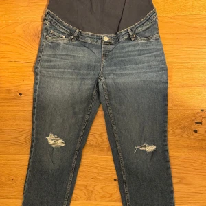 Blå mammajeans med slitningar - Snygga blå mammajeans från H&M med hög midja och rak passform vid ankeln. Jeansen har coola slitningar på båda benen och en mjuk, stretchig mudd över magen för extra komfort. Perfekta för dig som vill ha både stil och bekvämlighet under graviditeten.