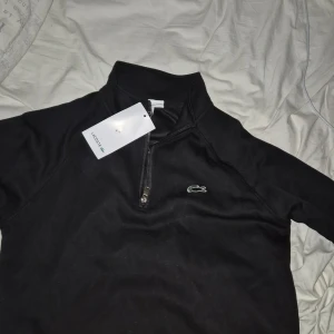 Svart zip från Lacoste stl s/xs - Svart långärmad tröja från Lacoste med klassisk ståkrage och half zip-dragkedja framtill. Tröjan har det ikoniska krokodilmärket broderat på bröstet och är i regular fit. Perfekt för dig som gillar stilren och sportig vibe.