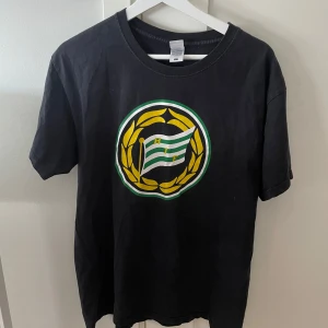 Svart Hammarby t-shirt med tryck - Svart t-shirt med stort Hammarby-tryck på bröstet i grönt, gult och vitt. Klassisk rund hals och korta ärmar. Tillverkad i mjuk bomull, perfekt för dig som vill visa ditt stöd för Bajen.