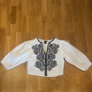 Vit blus med svart broderi från Lindex - Superfin vit blus från Lindex med svarta broderade blommor och blad framtill. Blusen har vida trekvartsärmar och är gjord i ett lätt, luftigt linne tyg som känns perfekt för både varmare dagar men även kyliga kvällar. Snyggt broderad detalj längs halsringningen och framtill. Är i storlek S men jag skulle säga att den snarare kanske är en Xs