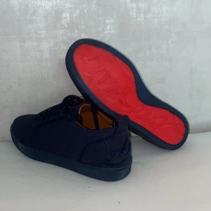 Mörkblå sneakers från Christian Louboutin - Exklusiva mörkblå sneakers från Christian Louboutin med klassisk röd sula och snygg broderad logga på hälen. Skorna har snörning, rund tå och är tillverkade i ett tygmaterial med läderdetaljer på insidan. Perfekta för dig som vill sticka ut med stil.