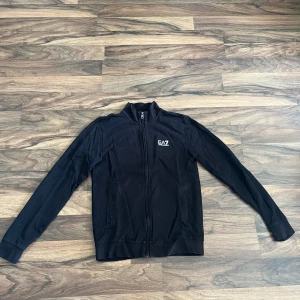 Svart Emporio Armani zipp - Svart Emporio Armani (EA7) med dragkedja hela vägen, ståkrage och två fickor framtill. Tröjan har en stilren look med EA7-logga på bröstet och är tillverkad i mjukt material. Skriv vid frågor!