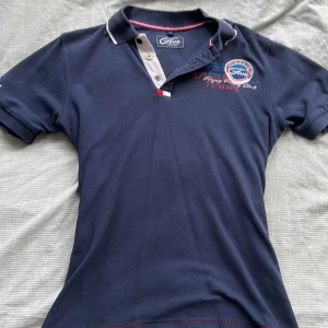 Mörkblå pikétröja från Five - Snygg mörkblå pikétröja från Five med broderade detaljer och emblem på bröstet och ärmen. Klassisk krage med kontrastkant och knappar framtill. Perfekt för en stilren och sportig look. Tillverkad i mjuk bomull och har korta ärmar.