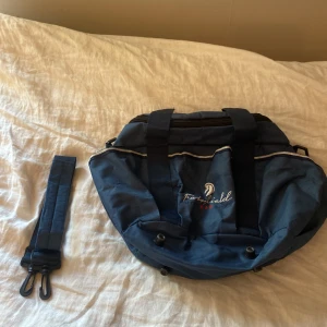 Blå axelväska från FairField - Säljer en blå axelväska från FairField med broderad logga och tre röda stjärnor framtill. Väskan har flera fack, avtagbar axelrem och praktiska handtag. Tillverkad i slitstark polyester och perfekt för dig som gillar att ha ordning på dina grejer.